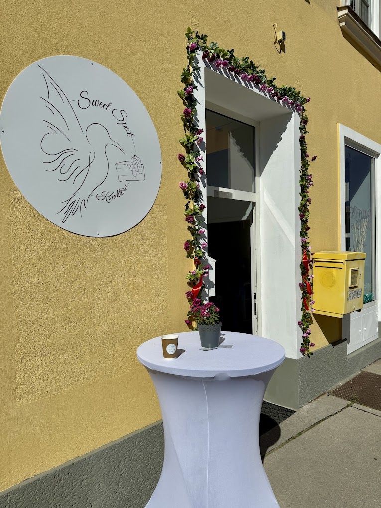 Eingang der Konditorei Sweet Spot mit rundem Logo-Schild an der gelben Fassade und blumengeschmückter Tür